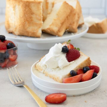 Gluten-free angel food cake slice with whipped cream and berries, cake behind.