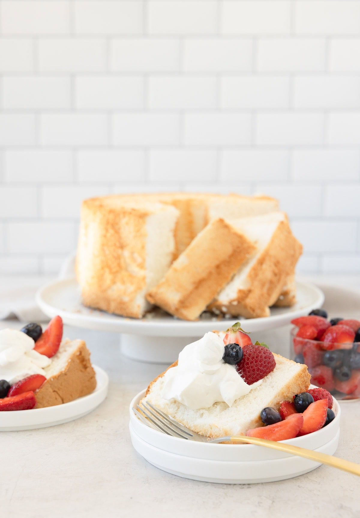 A slice of angel food cake topped with whipped cream, berries on the side, cake behind.