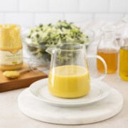 Creamy dijon mustard vinaigrette in a glass pitcher with mustard nearby and salad greens.