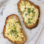 Open faced egg salad sandwiches topped with capers and herbs on a marble server.