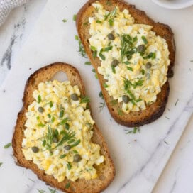 Open faced egg salad sandwiches topped with capers and herbs on a marble server.