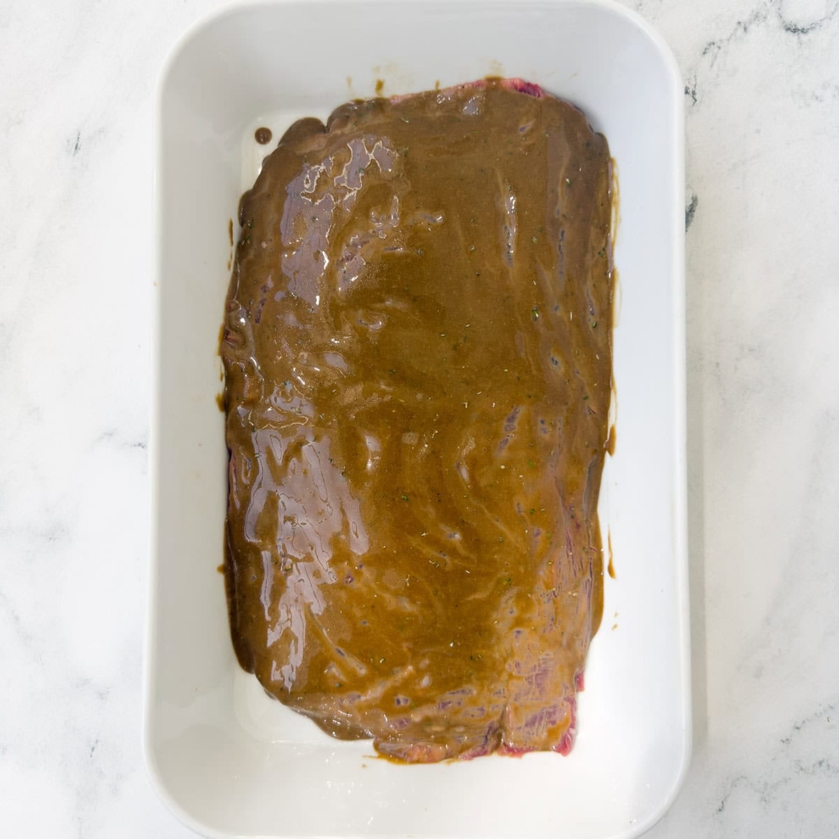 Steak marinating in a white rectangular casserole dish.