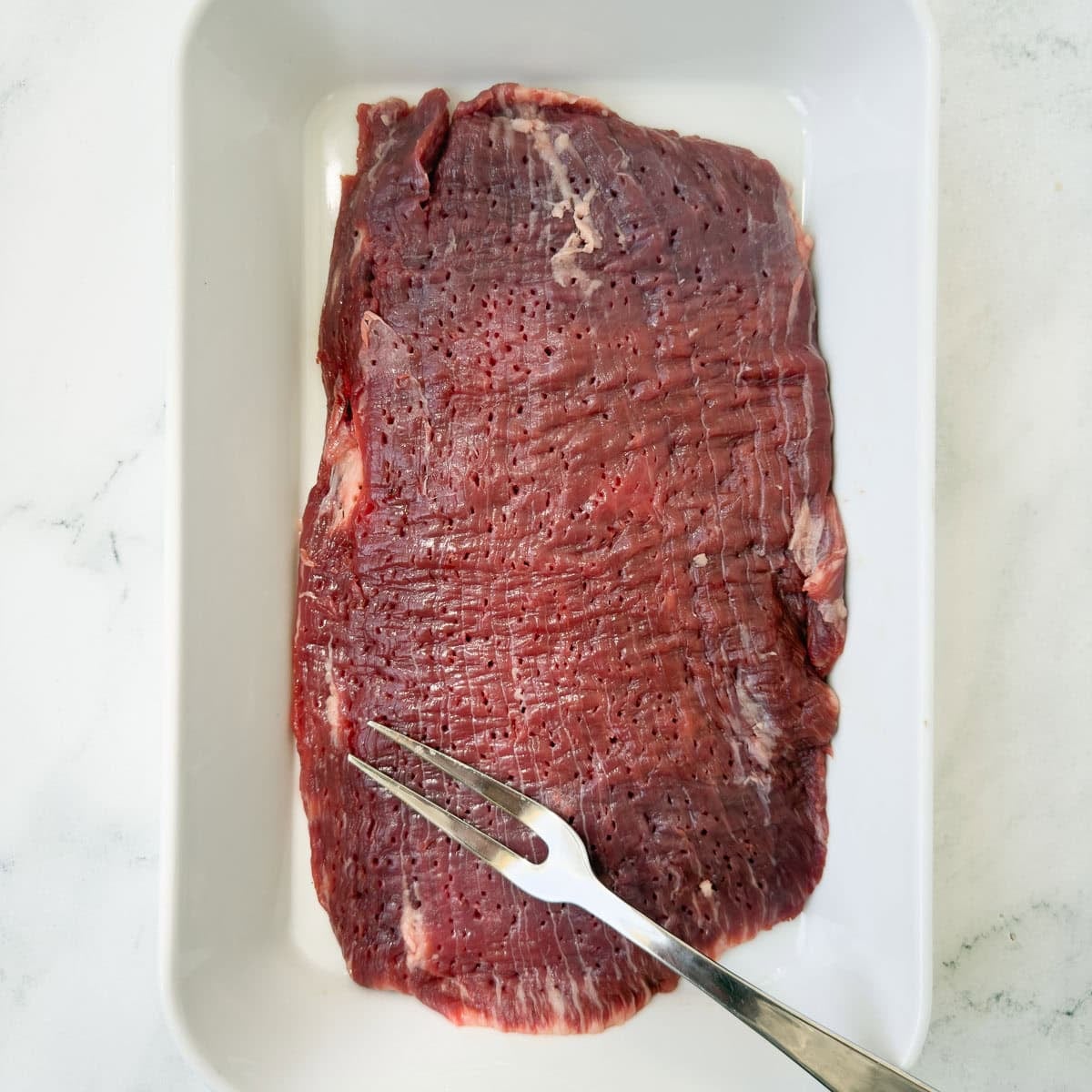 A flank steak in a white casserole dish with a meat fork.