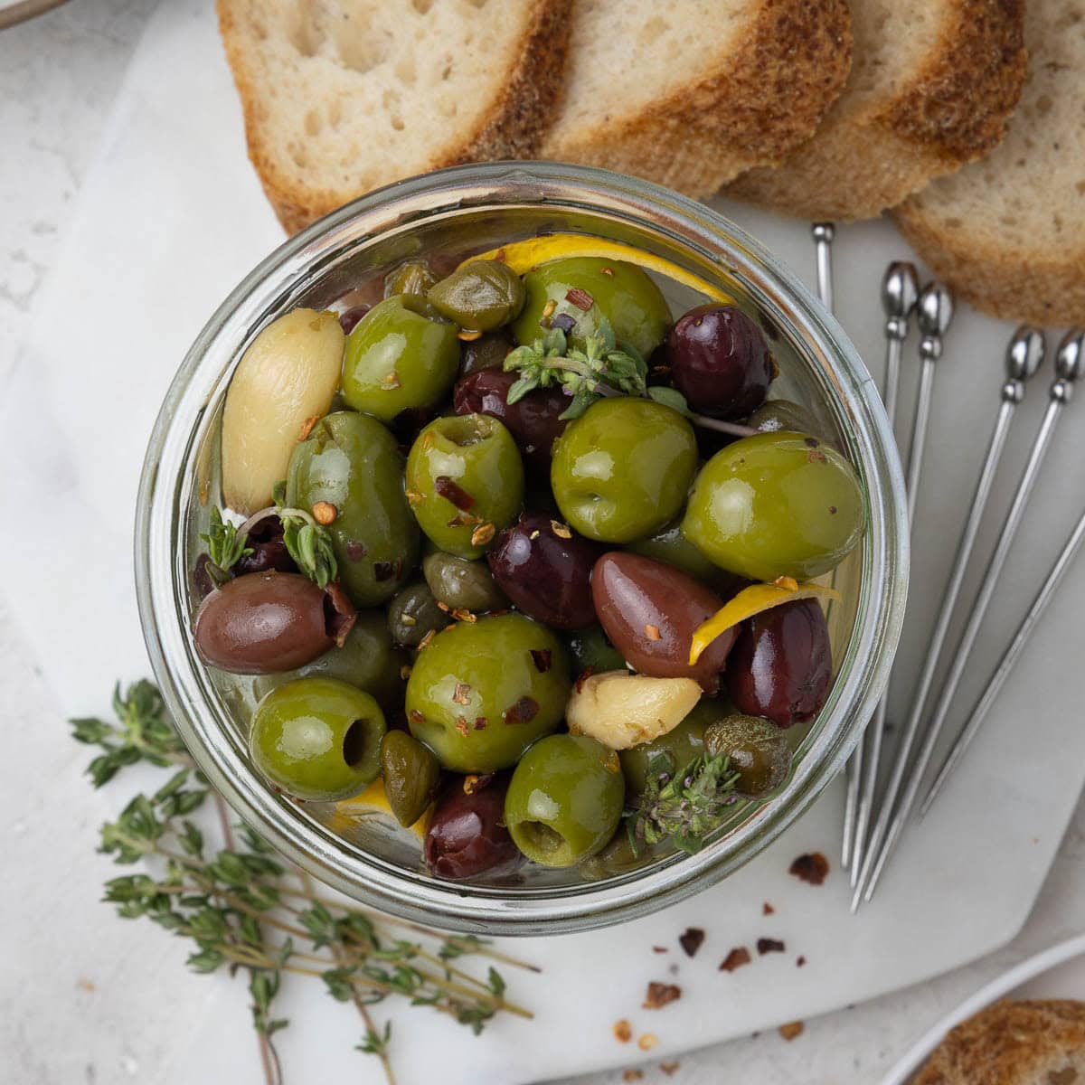 A jar of marinated olives with thyme sprigs, bread slices on a marble board.