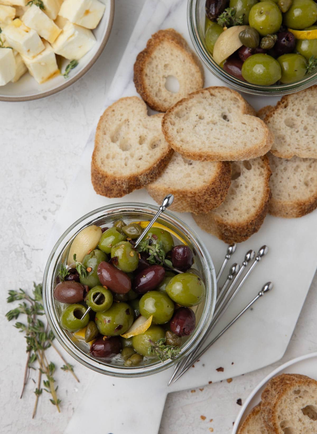 Jars of  msrinated black and green olives with thyme, baguettes slices, and feta cheese cubes.
