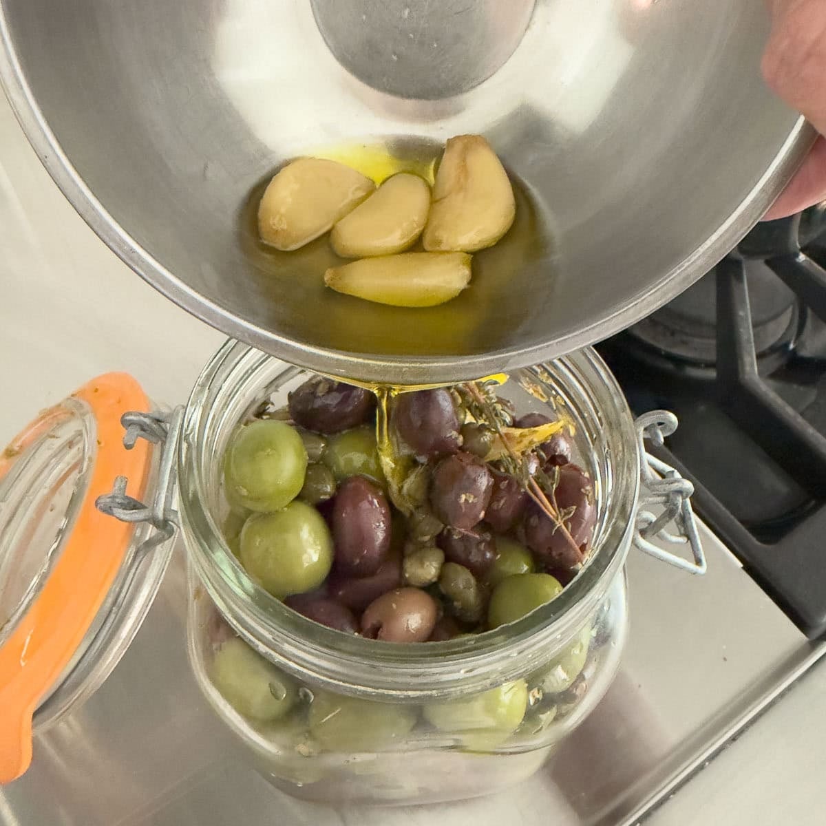 Pouring olive oil and garlic into a jar of olives for making marinated olives.