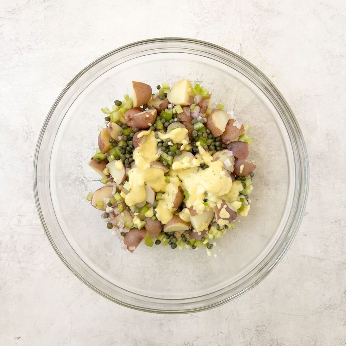 Dressing added to a red potato salad with celery, herbs, and capers.