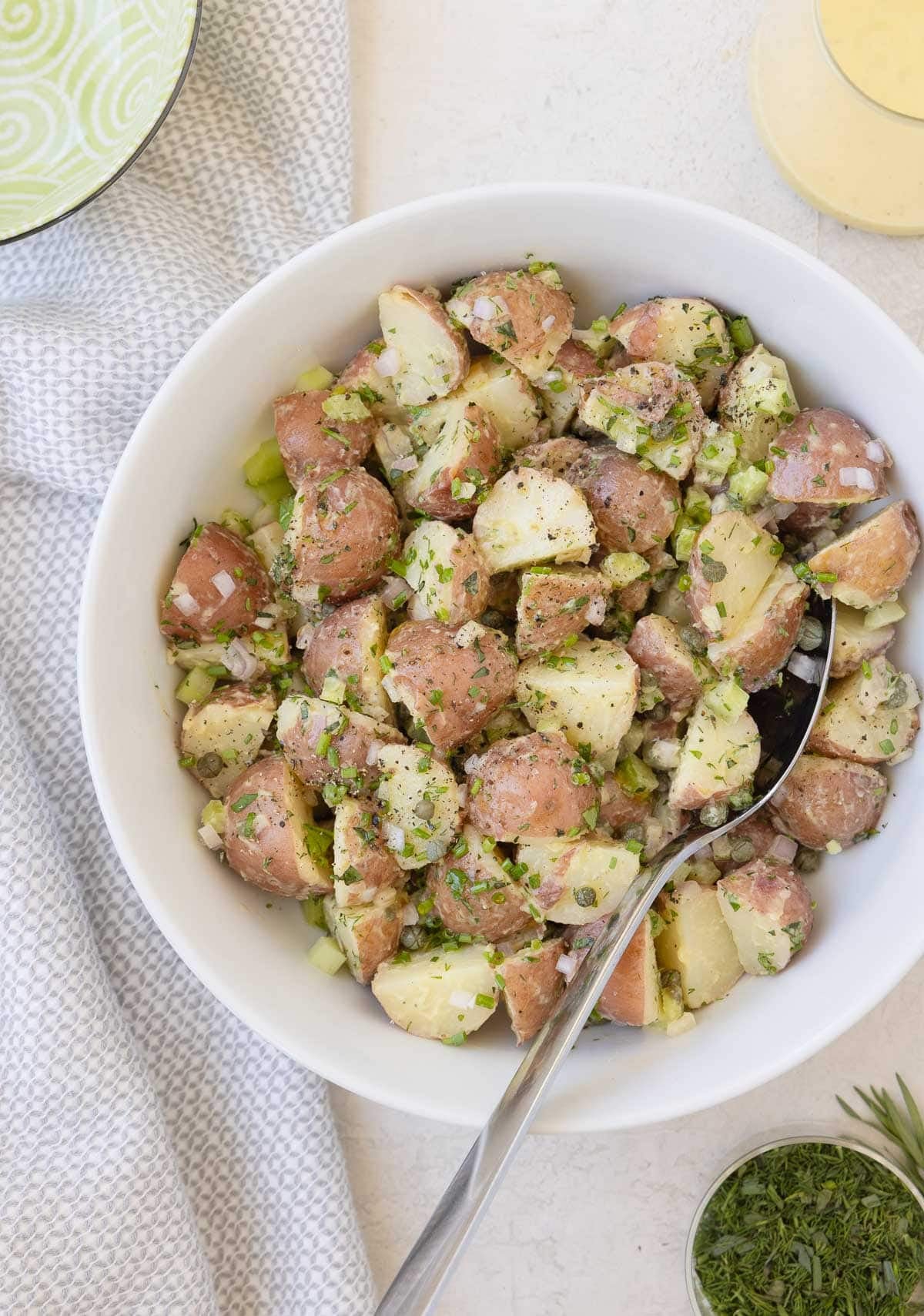 A white bowl of red potato salad with herbs, dressing pitcher, and a serving spoon.