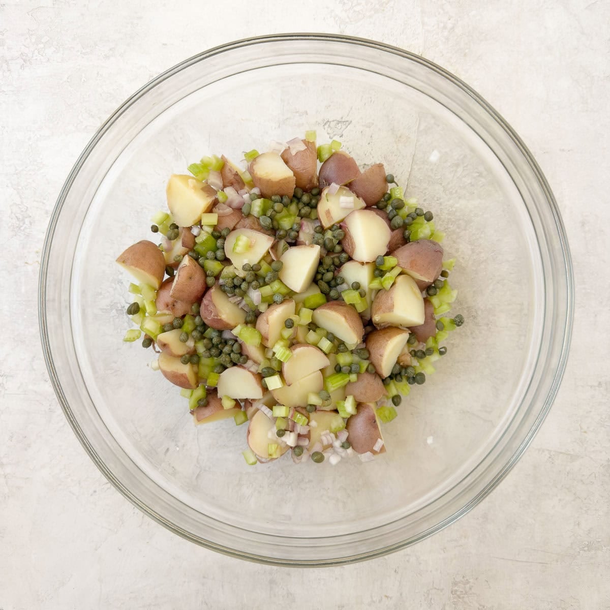 Red potato salad with celery, capers, herbs, and shallot in a glass bowl.
