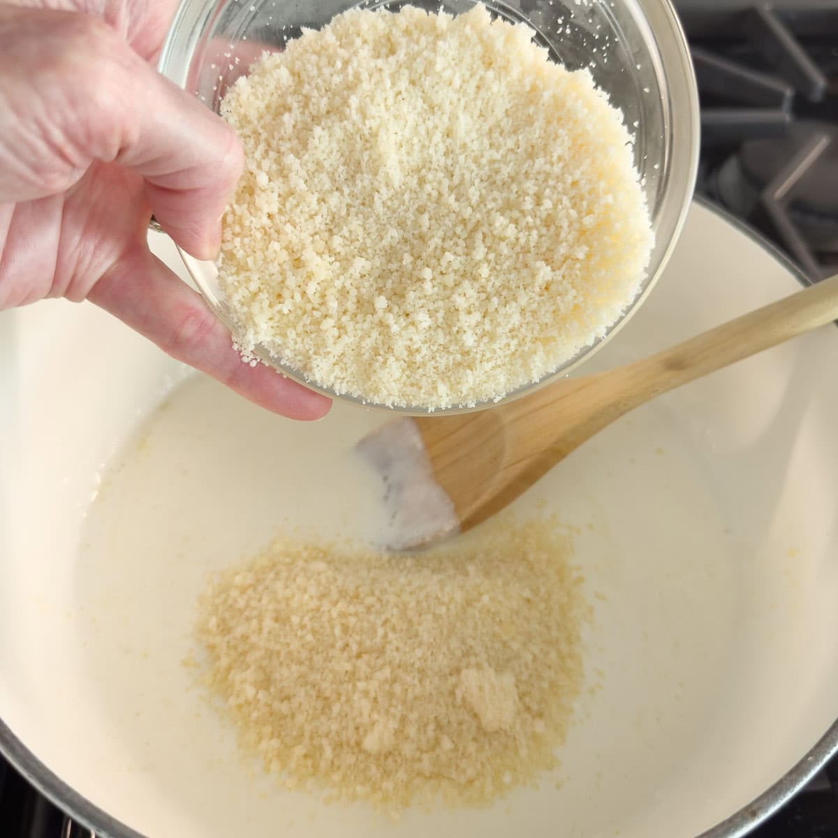 A bowl of grated parmesan cheese pouring into a pot for a cheese sauce.