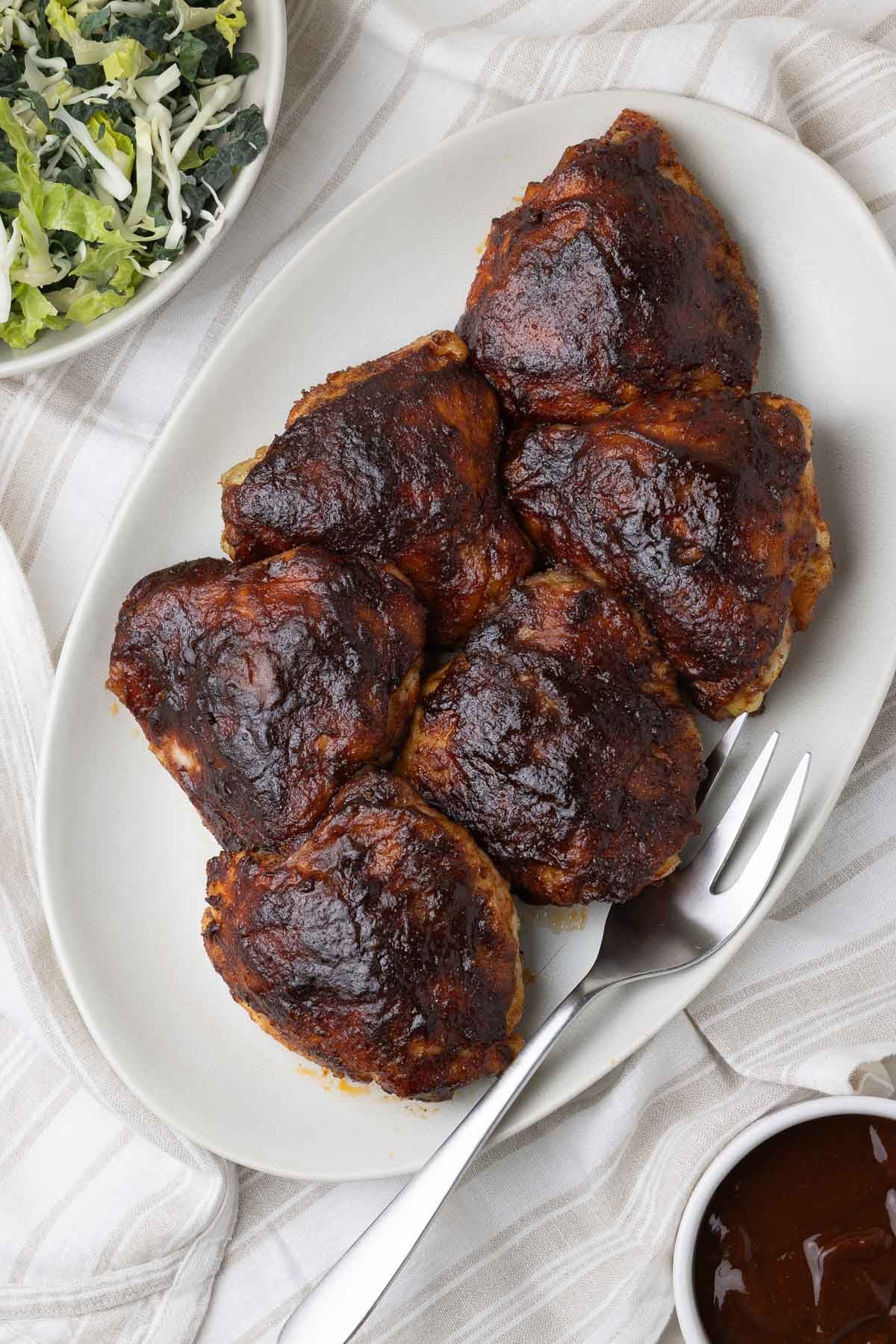 Oval platter of smoked chicken thighs glazed with bbq sauce and a meat fork on the side.