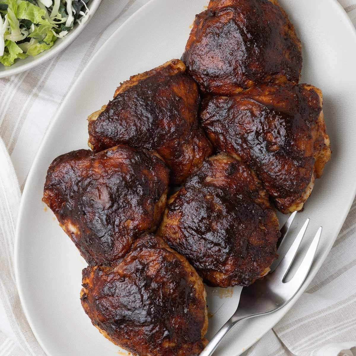 A platter of smoked chicken thighs with a dark glossy bbq glaze and salad on the side.