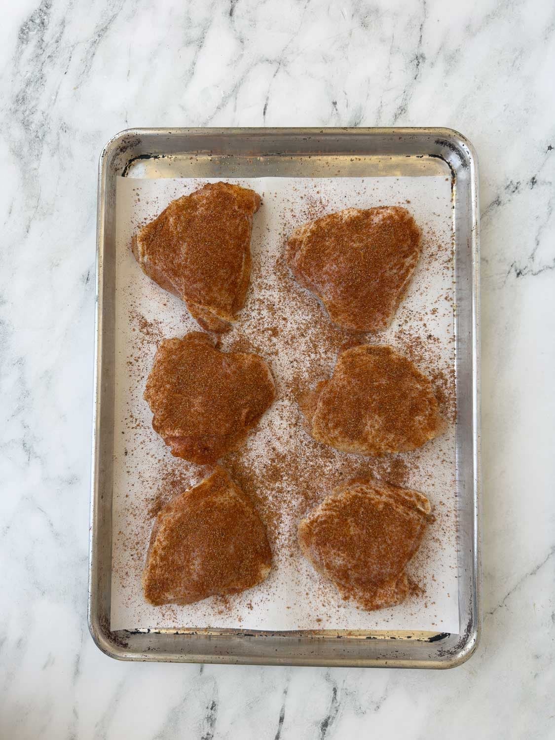 A rimmed baking sheet of seasoned bine-in chicken thighs on parchment paper.