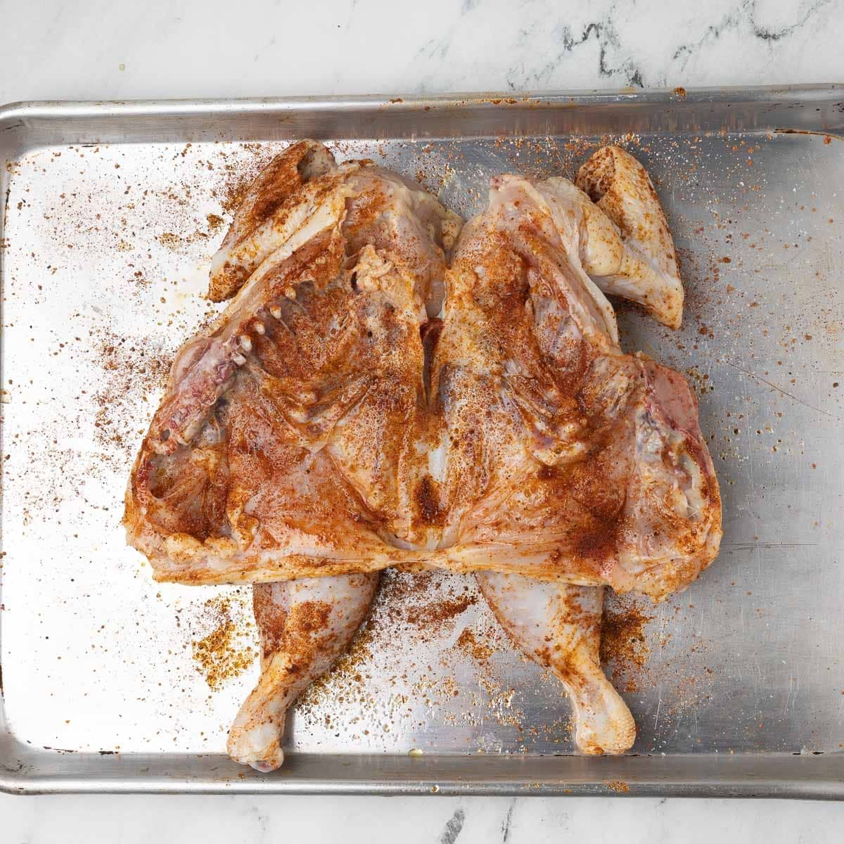 A spatchcocked chicken turned under side up with seasoning on a sheet tray.