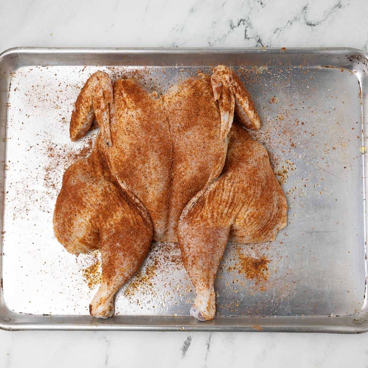 A dry rubbed spatchcock chicken ready for the grill on a rimmed baking sheet.