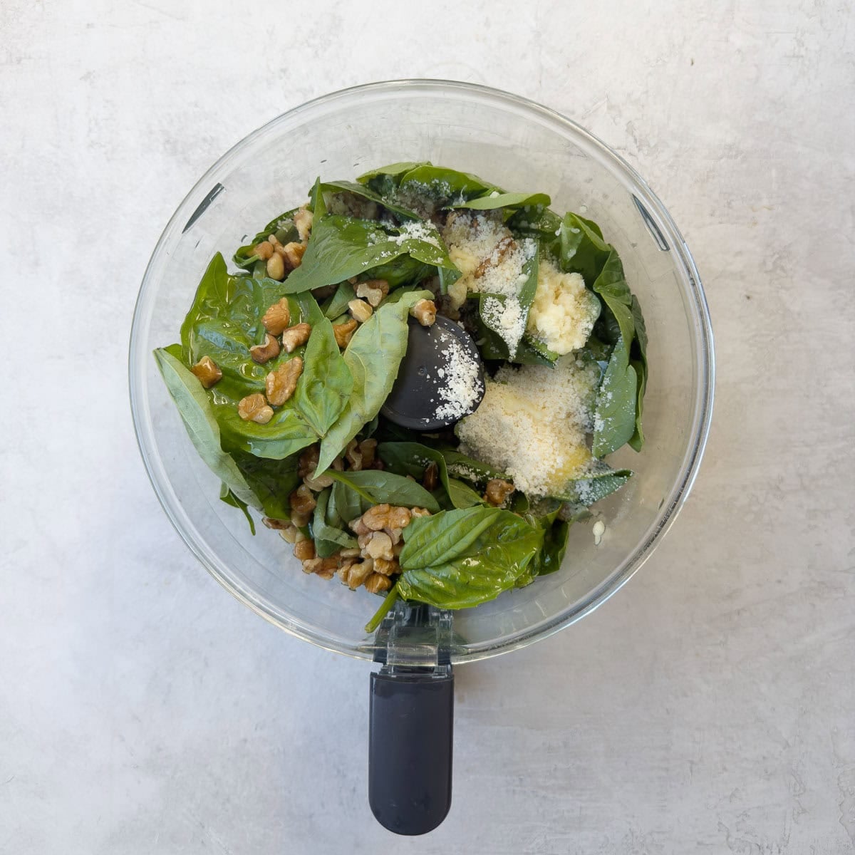Spinach and basil leaves, walnuts, parmesan, garlic, and lemon juice in a food processor workbowl to make pesto.