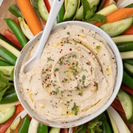 A bowl of cream white bean dip tipped with olive oil, pepper, and herbs with colorful vegetables all around.