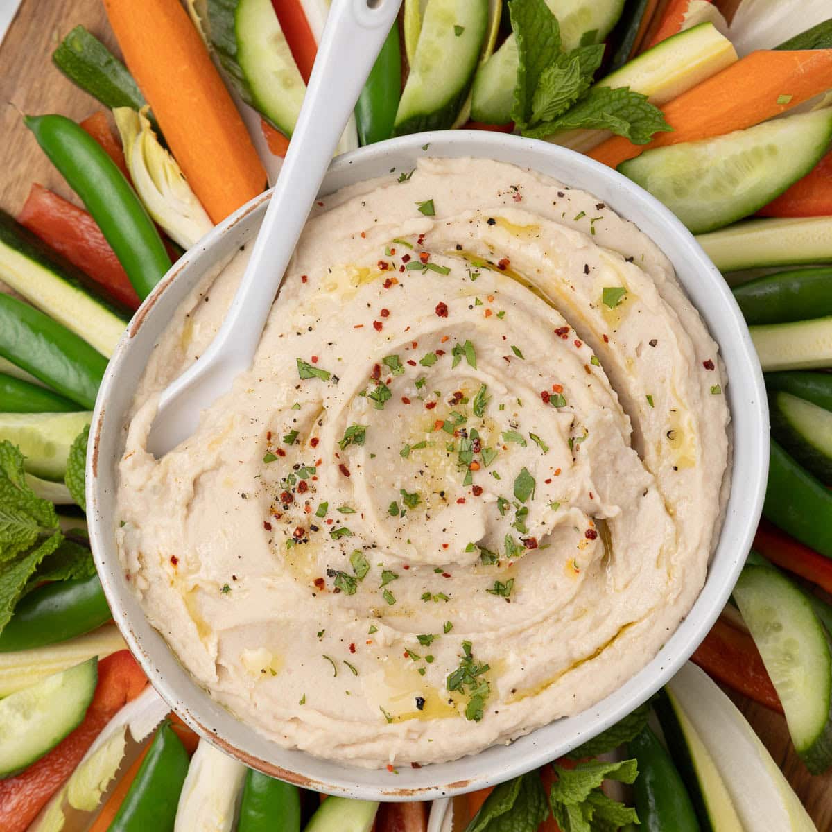 A bowl of cream white bean dip tipped with olive oil, pepper, and herbs with colorful vegetables all around.