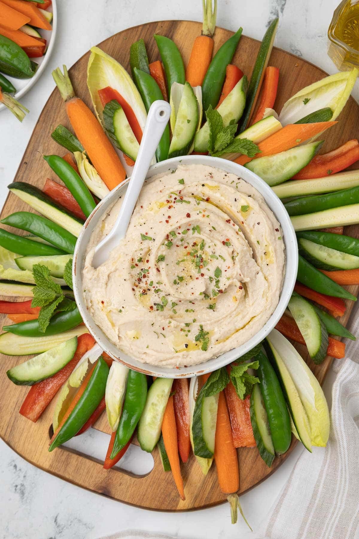 White bean dip drizzled with olive oil and sprinkled with herbs and pepper, with raw veggies.
