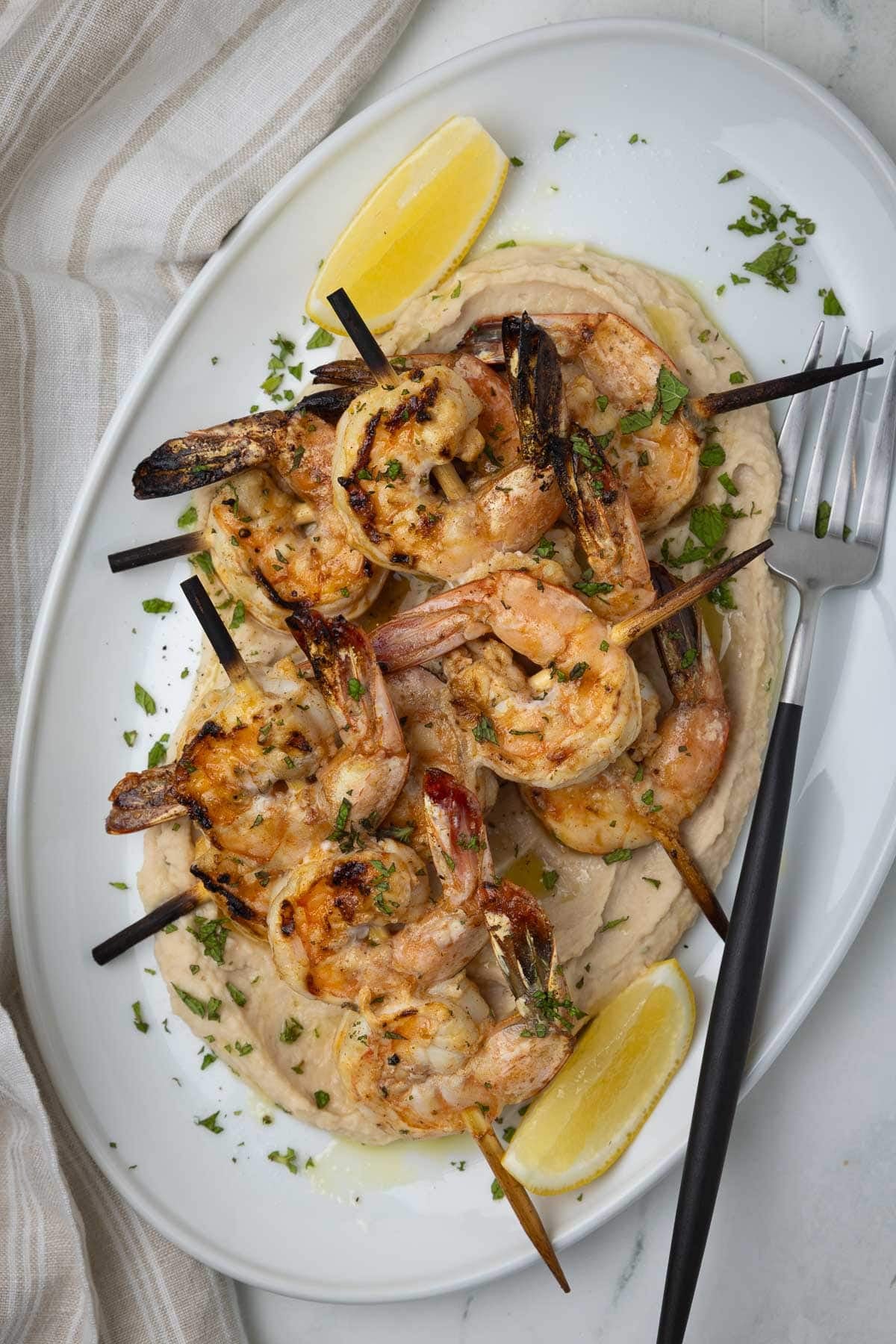 A platter of grilled shrimp atop white bean puree with lemon wedges and herbs.