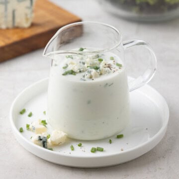 Yogurt blue cheese dressing with chuves and blue cheese crumbles in a glass pitcher with salad greens.
