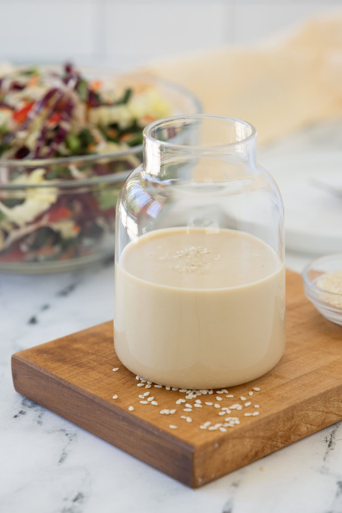 A glass jar of creamy beige sesame ginger dressing with white sesame seeds and a salad behind.