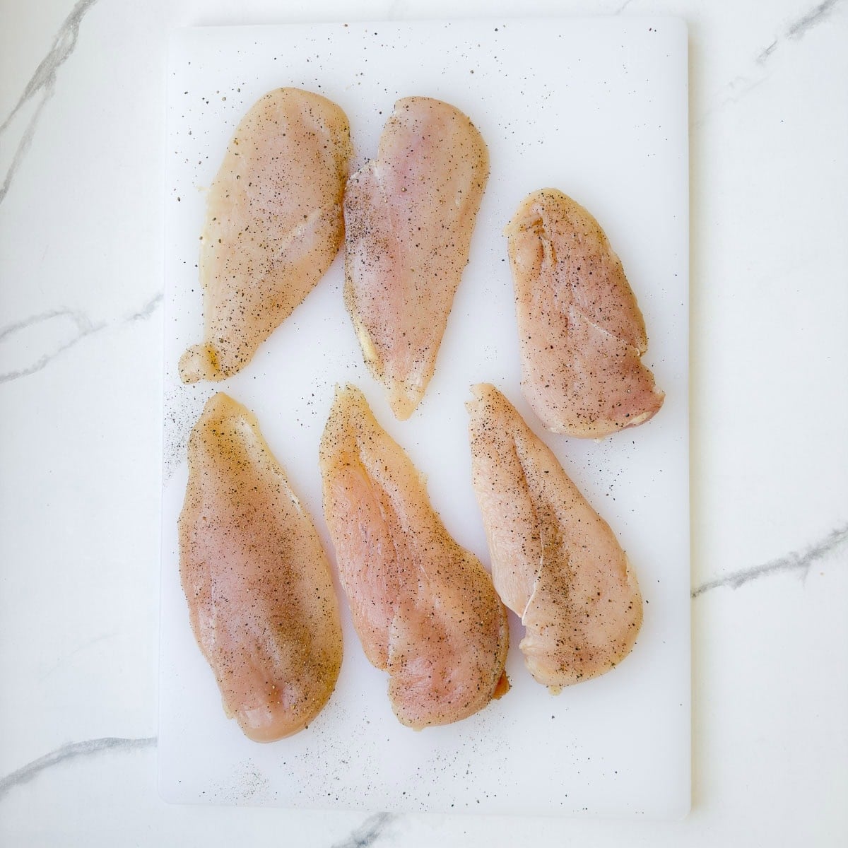 Split boneless skinless chicken breasts seasoned on a white cutting board.
