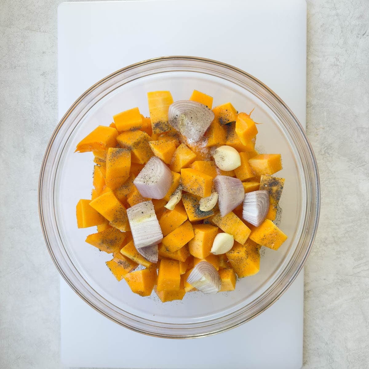 Cubed butternut squash, garlic clovers, and shopped shallots in a bowl tossed with oil.