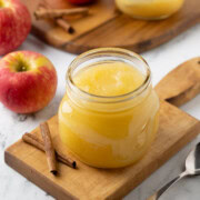 A jar or homemade applesauce with apples on the side, and cinnamon sticks.