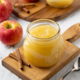 A jar or homemade applesauce with apples on the side, and cinnamon sticks.