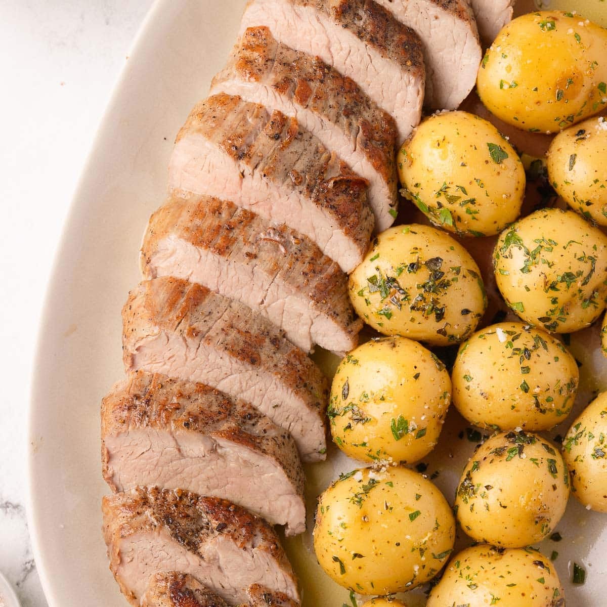 A serving plate filled with sliced oven roasted pork tenderloin and gold baby potatoes with herbs.