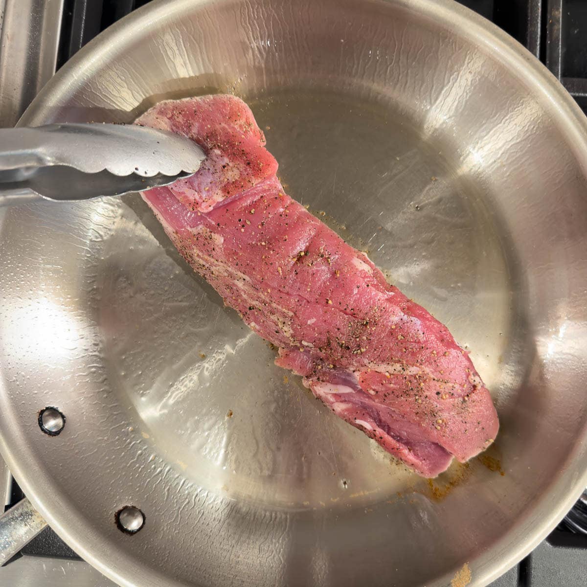 Searing pork tenderloin in a hot skillet, with tongs.